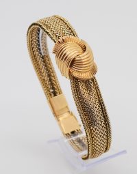 Gold-toned braided bracelet with a sculpted knot centerpiece on a clear display stand.
