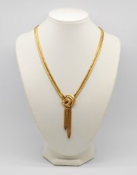 Gold tone chain necklace with a knot and tassel pendant displayed on a white bust stand.