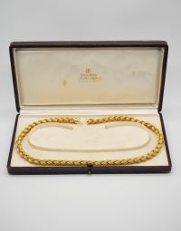 Gold braided necklace laid in an open velvet jewelry box with a light interior and a small logo on the lid interior.