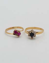 Two gold rings on a white background: left ring with two purple gemstones, right ring with a black center stone surrounded by small clear stones.
