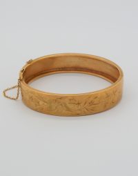 Gold-toned engraved bangle bracelet with a floral/leaf pattern and a small safety chain