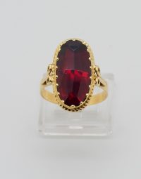 Gold ring with an oval deep red gemstone set in a beaded bezel, on a clear display stand.